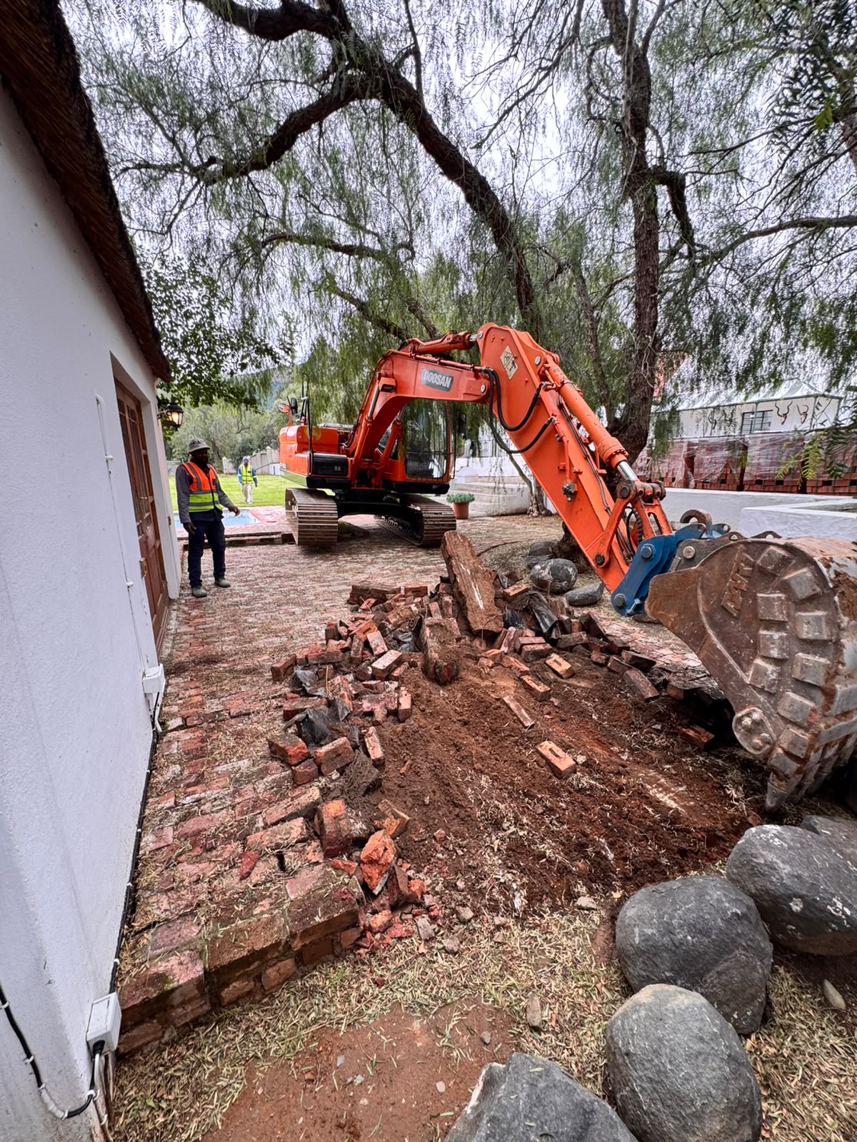 Excavator lifting old brick paving beside a house